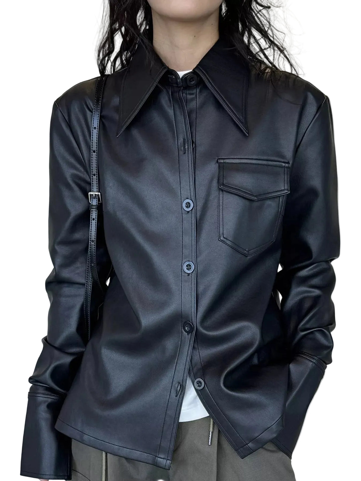 A person wearing a black faux-leather button-up shirt jacket with a pointed collar and a chest pocket, paired with gray pants and a black shoulder bag, set against a neutral cream curtain background.
