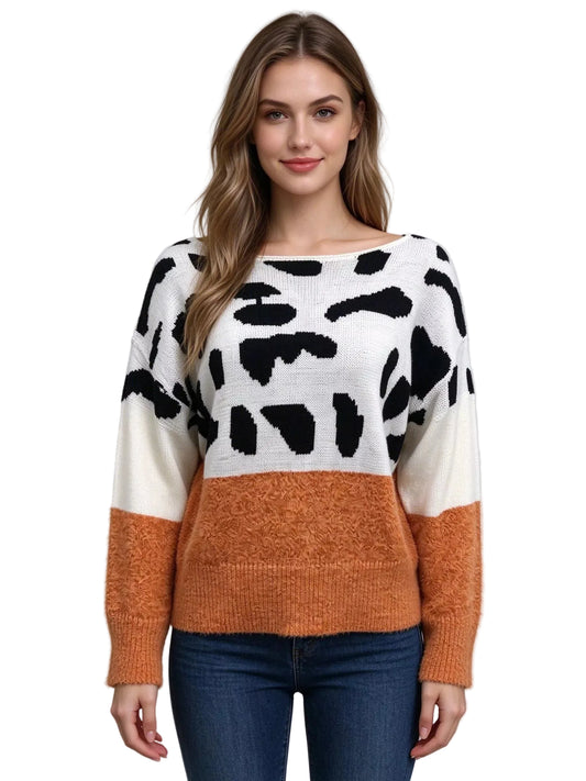 A woman modeling a color-block knit sweater with a black and white cow-print pattern on the upper section, solid white sleeves, and a textured rust-orange lower section and cuffs, paired with blue jeans against a light gray studio background.
