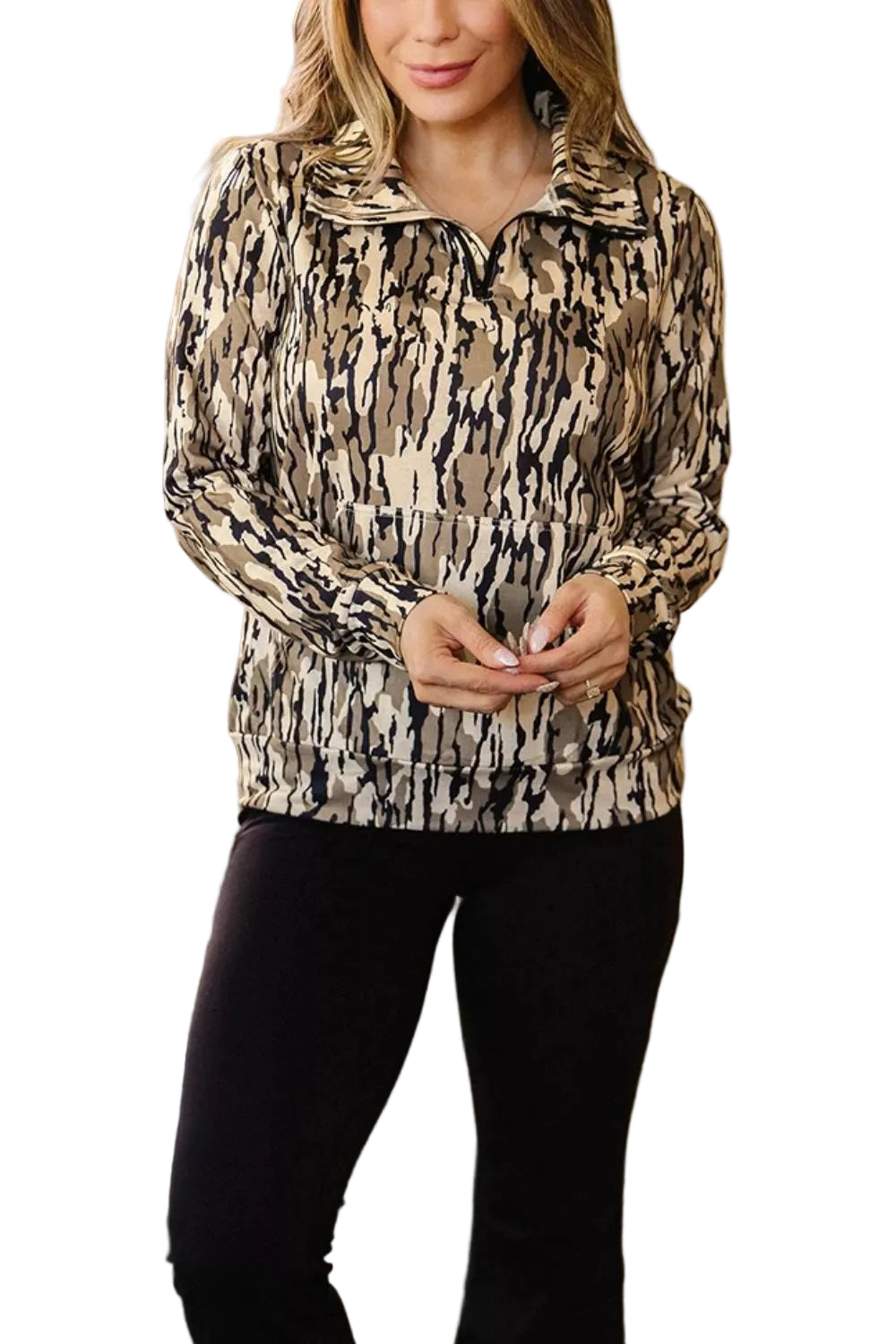 A woman wearing a beige, black, and brown abstract animal-print half-zip pullover jacket with black pants, standing in a room with a green couch in the background.