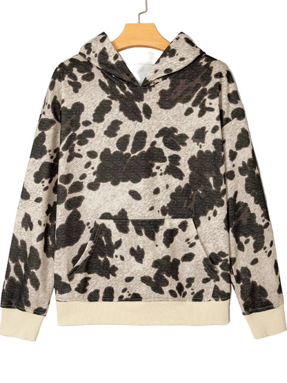 A long-sleeve hooded sweatshirt with a cow-print pattern in black and beige, hanging on a wooden hanger against a light gray textured background.