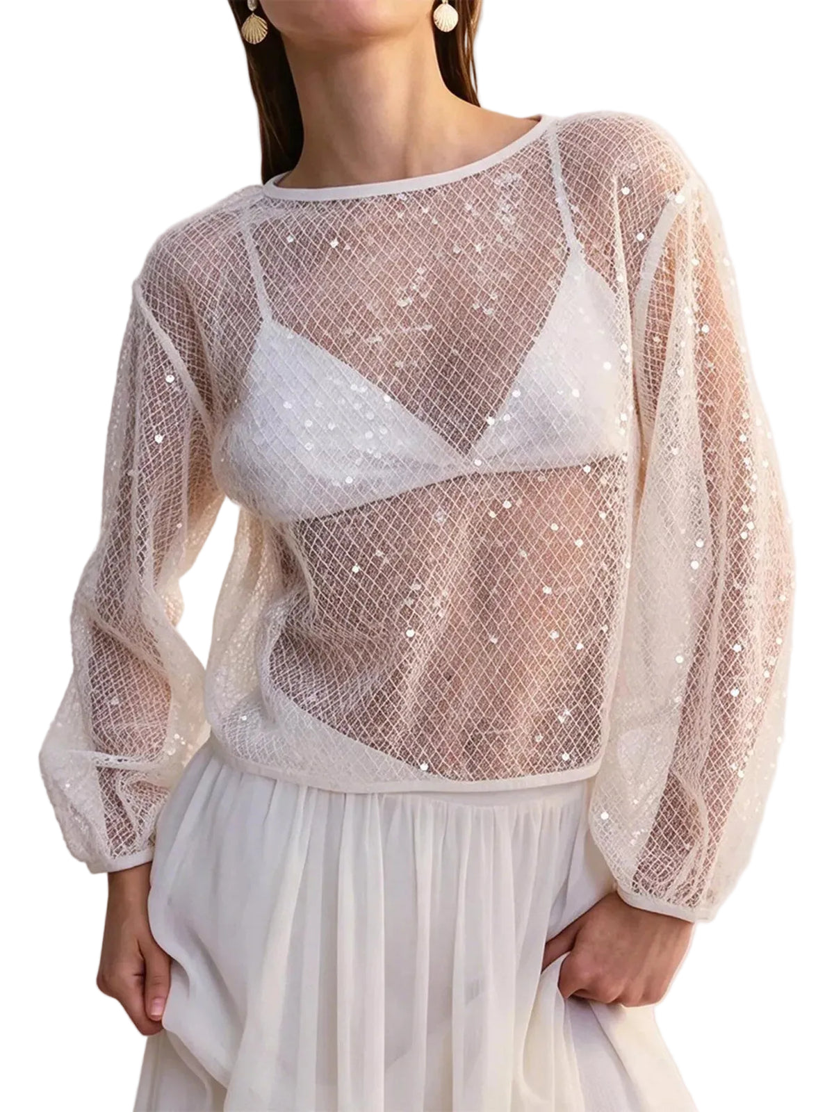 A woman wearing a sheer white mesh long-sleeve top with sequins over a white bralette, paired with a white skirt, against a plain light background.