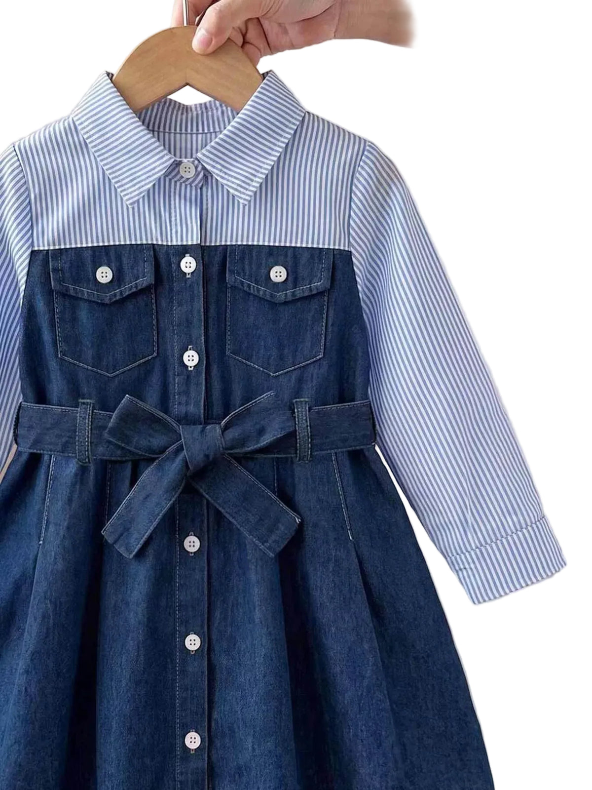 A children's dress hanging on a wooden hanger, featuring a blue and white striped long-sleeve shirt bodice with a pointed collar and button front, combined with a dark blue denim-style pinafore front panel, chest pockets, and a matching waist tie. A hand is holding the hanger, and part of a white dress is visible on the right.