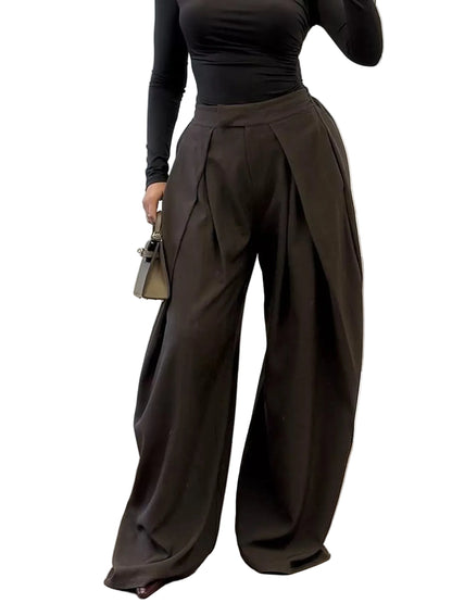 A person wearing high-waisted, wide-leg pleated pants in a dark brown color, paired with a black long-sleeve top, standing in a modern indoor setting.