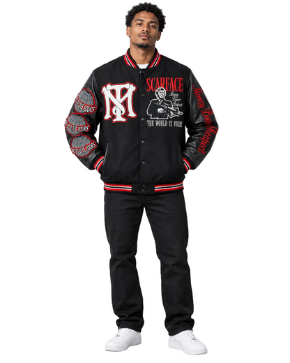 A man modeling a black varsity-style jacket with red and white striped ribbed trim, featuring a large red and white 'M' logo on the chest, 'SCARFACE' text, and graphic prints on the sleeves, paired with black pants and white sneakers against a light gray studio background.