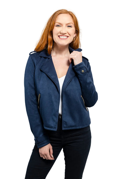 A woman with red hair wearing a navy blue suede moto jacket, a white top, and black pants, posing against a light gray studio background.