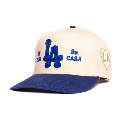 A baseball cap with an off-white crown and a navy blue visor, featuring a large blue embroidered 'LA' logo, the text 'Su CASA' and 'No CASA' in blue, and a gold heart outline with 'LA' on the side.