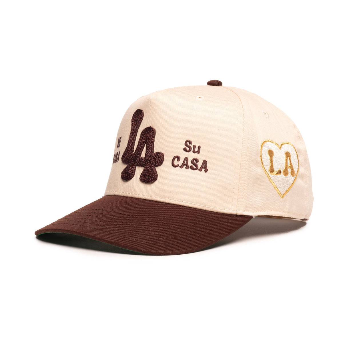 A baseball cap with a cream-colored crown and a dark brown visor, featuring embroidered details including a brown 'LA' on the front, 'Su CASA' text, and a gold heart-shaped 'LA' patch on the side.