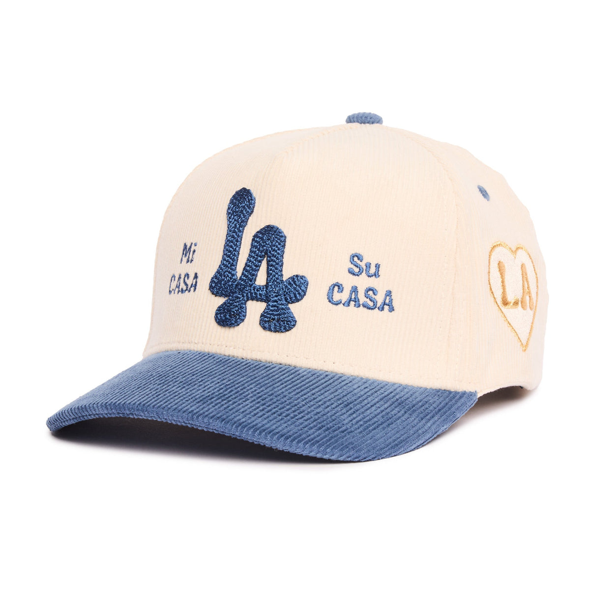 A baseball cap with a cream-colored crown, a blue denim-look visor, and blue embroidered details including a large 'LA' logo and the Spanish text 'Mi Casa Su Casa'. A gold heart outline with 'LA' is embroidered on the side.