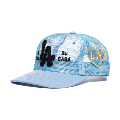 A light blue trucker-style cap with a mesh back, featuring a black embroidered 'LA' on the front, 'Su CASA' text, and a gold-outlined heart patch with 'LA' on the side.