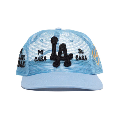 A light blue mesh-back trucker cap with a black embroidered 'LA' logo on the front, 'Mi Casa Su Casa' text on the sides, and yellow accents on the side panels, shown against a plain white background.