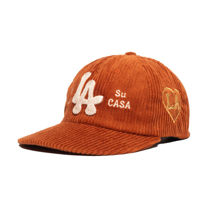 An orange-brown corduroy baseball cap with white embroidered 'LA' and 'Su CASA' text on the front, and a gold heart-shaped 'LA' patch on the side, shown on a plain white background.