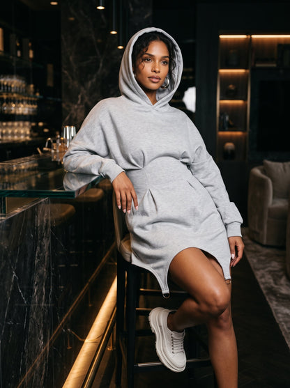 A woman wearing a light gray oversized hoodie dress sits on a stool in a dimly lit, modern bar or lounge setting. The dress has a hood, long sleeves, and a relaxed fit, paired with white sneakers.
